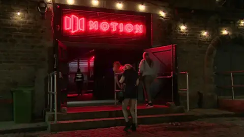 The entrance of Motion, with the word in red lights and a logo next to it. Women are walking in through the doors and it is night time.