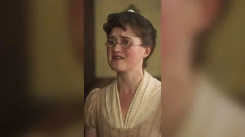 A young woman with brown hair looks distressed. She has rounded glasses and is wearing an old-fashioned cream coloured dress.