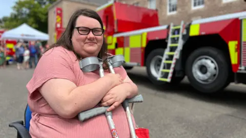 Vicky smiles at the camera as she sits in a chair holding her crutches. Behind he is a fire engine and crowds looking at stalls. She is wearing a pink dress.