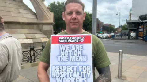 Ross Lopes-Lister has short fair hair swept back from his forehead and is wearing a green t-shirt. His left arm is heavily tattooed. He is holding a sign  saying "we are not on the menu, respect for hospitality workers".     