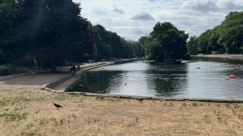 LDRS A view of a park, with a boating lake surrounding by a paved path. A grassy areas is dry and yellowing.