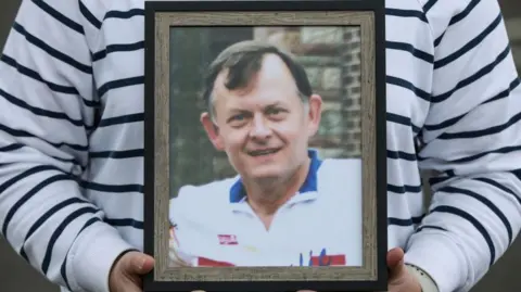A photo of Sean Brown being held up in a woman's hands. Sean Brown has dark hair and is wearing a white polo shirt. It is in a brown frame and the woman holding it up is wearing a white jumper with thin navy stripes. Only her torso and hands are visible.
