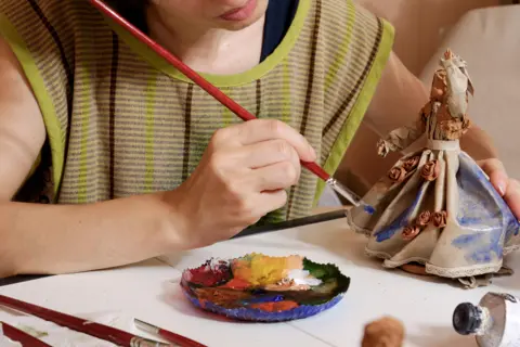 Getty Images An artist paints a figurine. She is holding a paintbrush in one hand and has a dish with paint in front of her on a covered table. The dish has different colours in it, including reds, yellows and white. The ceramic figurine is partly painted with her hand raised and has a long flowing dress.