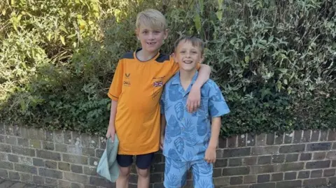 Two young boys standing side-by-side. A blonde boy in an orange shirt has his arm around a shorter boy with brown hair who is wearing a blue shirt.