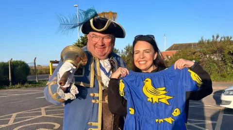 Bob Smitherman, wearing the tricorn hat and ceremonial robes of the Worthing Town Crier, stands with his bell on his shoulder, while Alison Ferns stands beside him, holding up a dress decorated with the Sussex coat of arms.