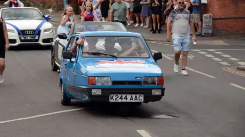 Erewash Borough Council A blue Reliant Robin driving down a road with a white Audi behind it. There are three people visible inside the Reliant Robin and a crowd of people to the right on the pavement.