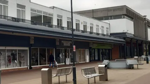 LDRS A row of retail units which are housed in a white 80s-style building. The high street has silver benches and bins outside.