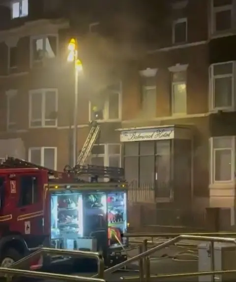Eddy Chapman/Bridlington Leisure Association A night-time street scene showing a brick multi-storey building with smoke coming from a first-floor window. A fire engine is parked in the foreground with an equipment compartment open and lit, showing hoses and tools inside. A ladder is extended from the fire engine up to a window, from where a firefighter is positioned inside. Streetlights cast a yellow glow on the building façade, and a sign above the entrance reads “Balmoral Hotel". Fire hoses run along the pavement in front of the building.