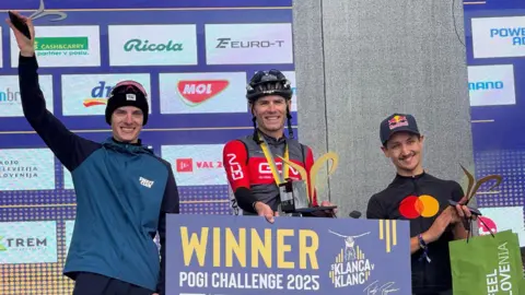 Georgia Basetta é retratada em Andrew Father Podium, ele mantém uma placa que está escrita "Vencedor, Desafio Pogi 2025"