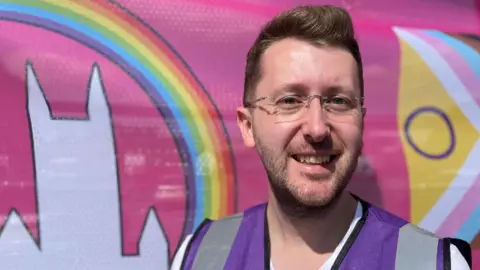 Arran Hart smiles as he stands in front of a pink Lincoln Pride banner, which features a rainbow flying over a white outline of Lincoln Cathedral. He is wearing a purple hi-vis vest and glasses.