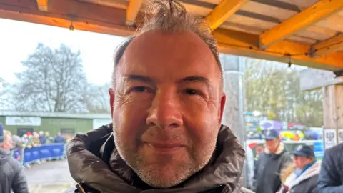 Marcus Stewart looks to the camera. He has thin grey hair and is wearing a black puffer jacket.