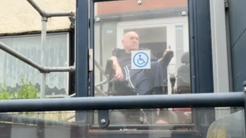 A man in a wheelchair looks out from behind a glass lift door outside a house