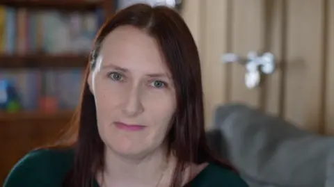 A woman with brown long hair looks straight at the camera in a head and shoulders image. She has green eyes and is wearing a green top. 