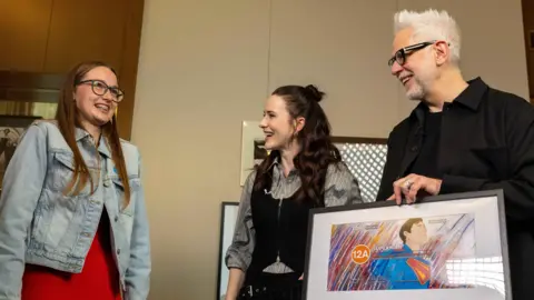 BBFC A girl in a denim jacket witha big smile as she talks to actor Rachel Brosnahan and director James Gunn in a room with soft, cream walls. James Gunn is holding a framed artwork depicting Superman in his blue superhero costume and an orange 12A age rating circle.