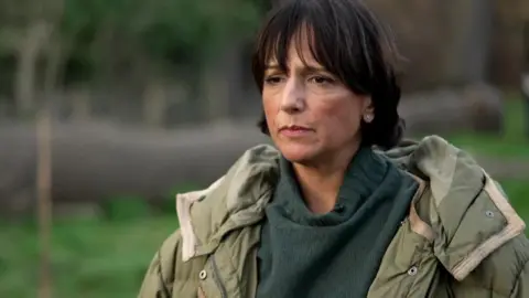 Head-and-shoulders image of a Romilla Arber wearing a green coat and jumper, standing outdoors in a park, with a young tree and blurred greenery visible in the background.