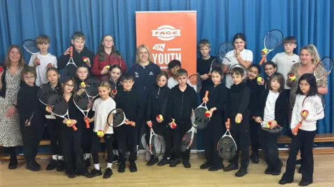 The image shows children posed with Sonay Kartal, a tennis player, in the middle of the photograph. They are all holding tennis rackets and tennis balls. Behind them is a blue curtain and an orange banner.