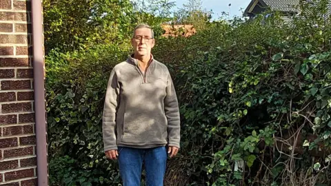 Local Democracy Reporting Service Paul Knights is standing between the wall of a house and a hedge. He is wearing a grey fleece jumper and blue jeans.