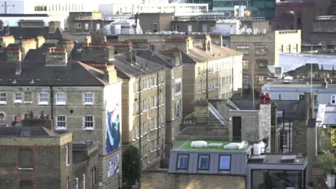 Brick residential buildings with rows of chimneys and rooftop terraces, viewed across a dense urban neighbourhood in daylight, with sunlight striking the façades and a painted mural visible on one wall.