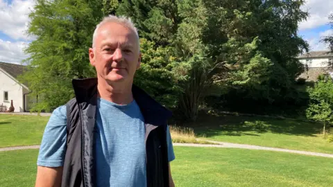 Chris Plumb is wearing a dark blue body warmer and a light blue T-shirt is standing in the foreground. He has short grey hair. In the background there is a large green tree and on the left there is a small white building.