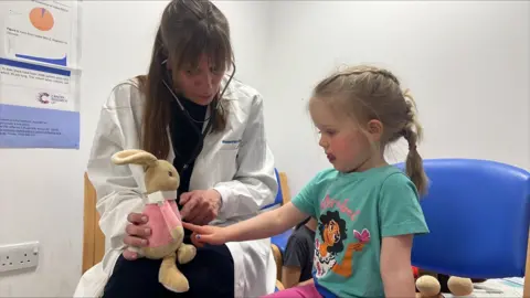 Richard Knights/BBC A young girl sits on a blue chair next to a doctor wearing a white lab coat. The doctor holds the girl's toy bunny on her lap while the girl touches the bunny's tummy, as if checking it over. The girl has a french plait in her hair and wears a turquoise Encanto t-shirt.