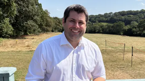 Leigh Frost wears a white shirt and has dark hair and stands in front of some parched lawns with trees in the distance 