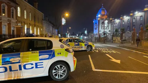 BBC Two police cars sit on an otherwise vehicle-free street at night time. Christmas decorations hang from lamp-posts, and two and three storey buildings of varying age and design face out onto the street.