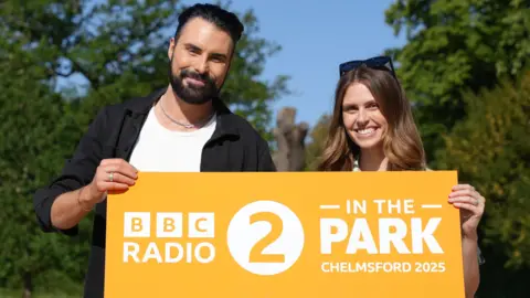 TV personality Rylan Clark and comedian Ellie Taylor holding a yellow banner that reads BBC Radio 2 in the Park Chelmsford 2025. They are both smiling at the camera. Ellie has sunglasses on her head.