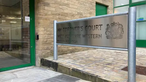 BBC Sign outside Bradford coroners court saying 'HM Coroner's Court West Yorkshire Western' with arrow and 'reception' on it. Door is partially in view.