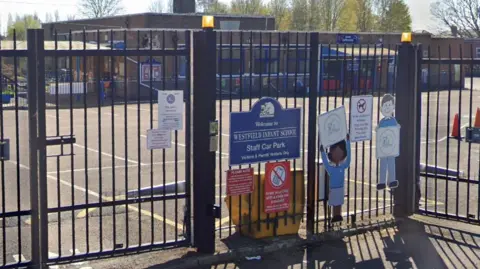 Google Outside of Westfield Infant School - a gate around the school is at the front of the photograph with signs on, and the playground and building is in the background. 