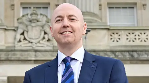 A man, Jon Burrows, is smiling at the camera. He is bald and is wearing a navy jacket over a white shirt and a blue and navy diagonally striped tie. He is standing outside of Parliament Buildings at Stormont in Belfast.
