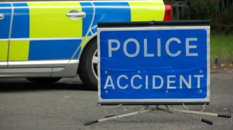 A sign reading POLICE ACCIDENT, with white letters on a blue background, that has been placed on a road. A police car is behind it.