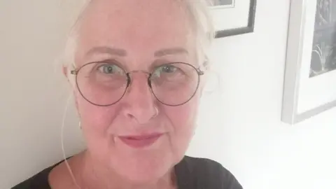 Sue Coleman A woman with white hair, tied back, wearing round black-rimmed spectacles and with a nose ring, smiles up at the camera as she takes a selfie. She is wearing a dark grey/black top and is standing against a white wall with framed pictures hanging on it.