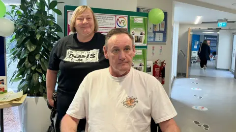 David Malpas, sat in a wheelchair with his wife Claire Malpas standing behind him in Torbay Hospital