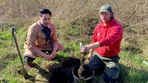 A man in a camel-coloured long coat crouching alongside another man wearing a red hooded top and holding a sapling in a black plant pot.