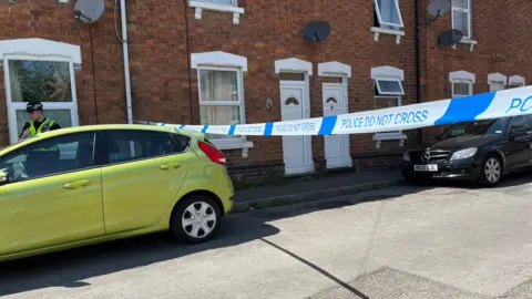BBC 'Police Do Not Cross' Tape creates a barrier across a street of terraced houses, with a police officer behind a green car parked on the street. A black car is also parked on the street.
