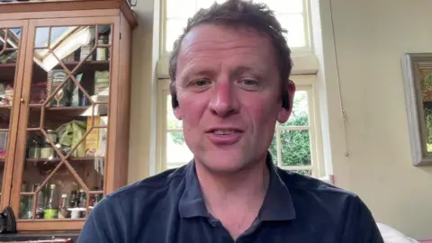 Christopher Wordsworth is sitting in a kitchen beside a window and pantry of jars doing a zoom interview. Only his head and shoulders are visible. He has brown hair and is wearing a blue top.