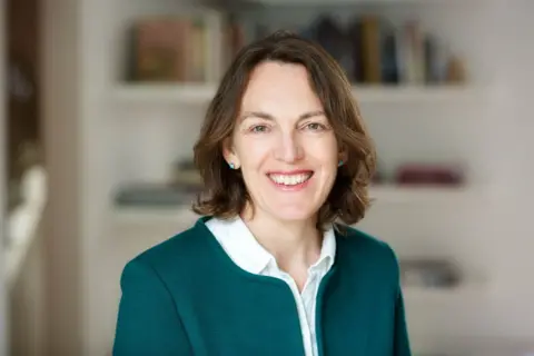 Louise Cooper Louise Cooper looking directly towards the camera, smiling. She is wearing a dark green blazer with a white shirt, with the top button unbuttoned. She has dark brown hair with some light patches .