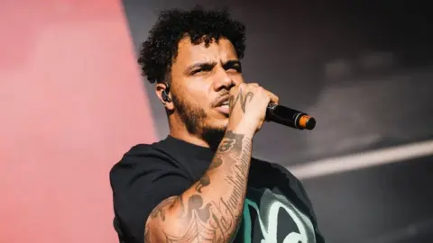 AJ Tracey performing at Laneway festival, he is wearing a black t-shirt and holding a microphone