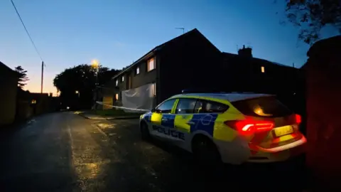 Police car with lights on outside a house, and police tape.