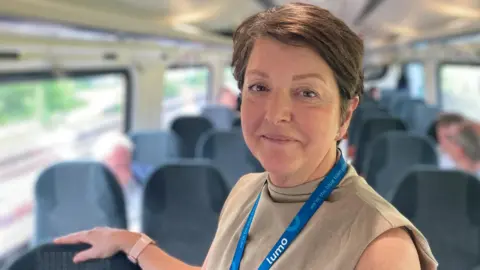 Close-up of Max Joicey. She is standing in a railway carriage with her right hand resting on the back of a seat. She is wearing a beige sleeveless top with a blue Lumo lanyard. She has short dark hair.