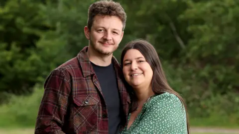 PA Media Thomas and Amy. Thomas has short dark hair and facial hair and Amy has long dark hair. They are standing outside with their arms are around each other and while looking at the camera and smiling.