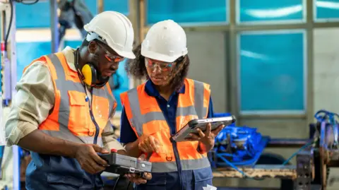 Two manufacturing process engineers on helmets and orange safety vets having a discussion about welding assembly process improvement to decrease parts defects during production in front of the the robot in industry factory. 