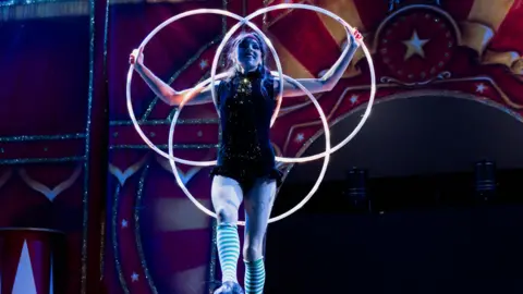 A circus performer holds up three hoops while smiling during a stage performance.