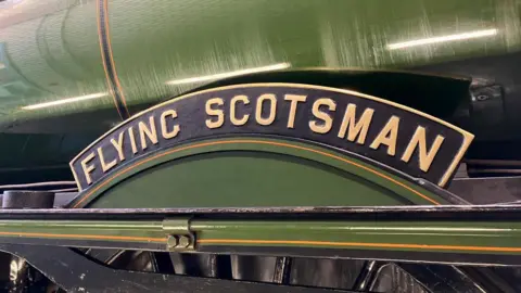 BBC A green-painted metal cylinder with a curved black sign which has the words flying Scotsman written on it in gold lettering and some black-painted metalwork below
