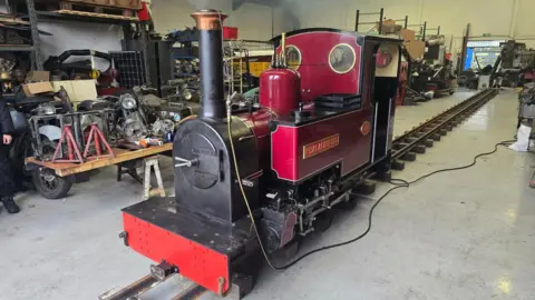 Joe Nemeth A red miniature steam locomotive sitting on some rails in a factory setting. Steam can be seen coming from the funnel