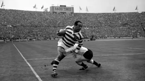 PA Media Billy Boston bursting through a tackle at a packed Wembley during the Rugby League Challenge Cup Final in 1959 in a black and white photo