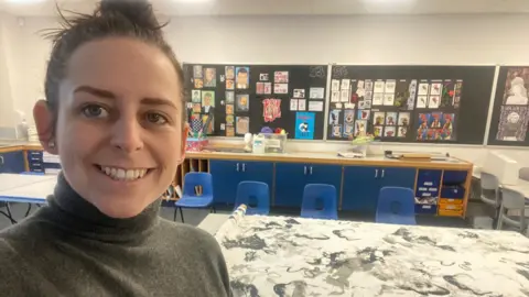 Nadia Pinkney taking a selfie in her art classroom. She is wearing a grey turtleneck jumper and her hair is in a bun. There are paintings on the wall and desks and chairs in the background.