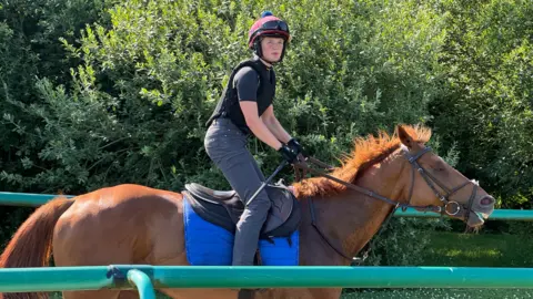 Lauren Frost in full riding gear standing in the stirrups as she canters a horse on the gallops. Shot is side on and shows green railings on either side.