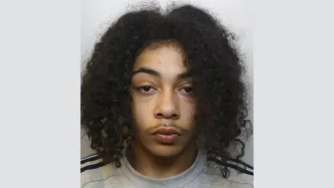 Avon and Somerset Police A police mugshot of Kodi Wescott. He has long dark curly hair, brown eyes and a thin moustache. He is wearing a grey sweatshirt with black stripes on the shoulders, and standing against a grey background. 