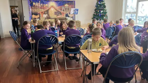 School children eating their lunch at Ingleton Primary School. The children are dressed in purple jumpers and are sitting at wooden desks. A Christmas tree stands at the far side of the room.